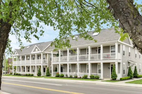 Two-storey apartment buildings in tan siding with white columned double porches, seen from across the road through overhanging branches.