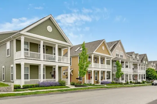 A street of two-storey homes in olive, gold and tan siding, each with white columned double porches and a clipped front lawn.